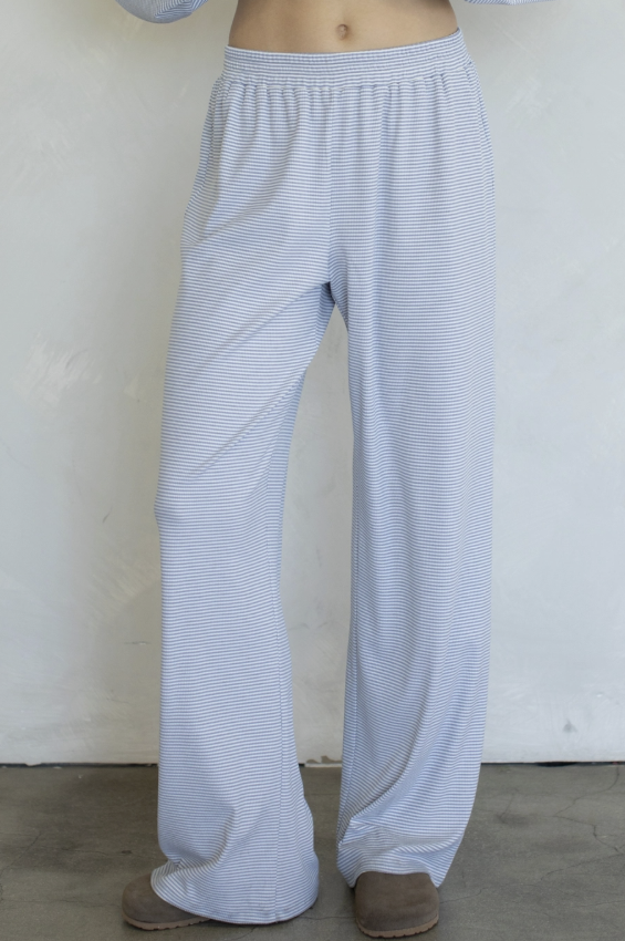 A person wearing light blue and white striped lounge pants with a wide elastic waistband and loose straight legs, standing against a plain off-white wall.
