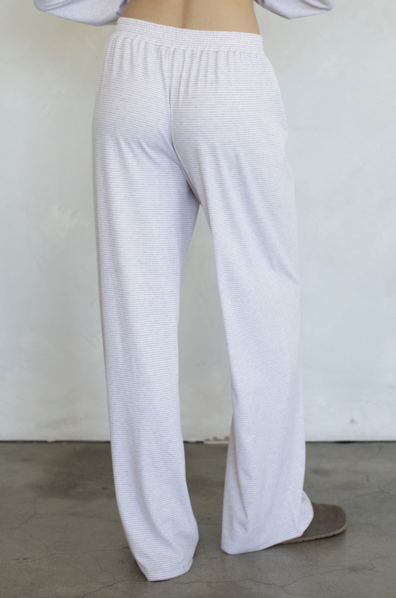 White textured lounge pants shown from the back on a model, featuring a wide elastic waistband and relaxed straight-leg fit, paired with a light long-sleeve top and a brown slip-on shoe.