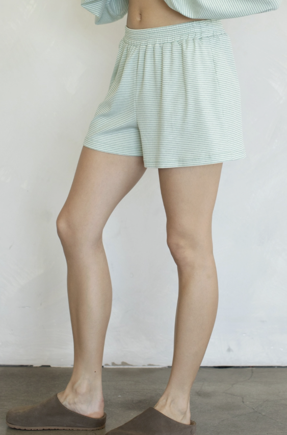 A person wearing light mint green and white striped lounge shorts, paired with brown slip-on slippers, standing against a white wall.