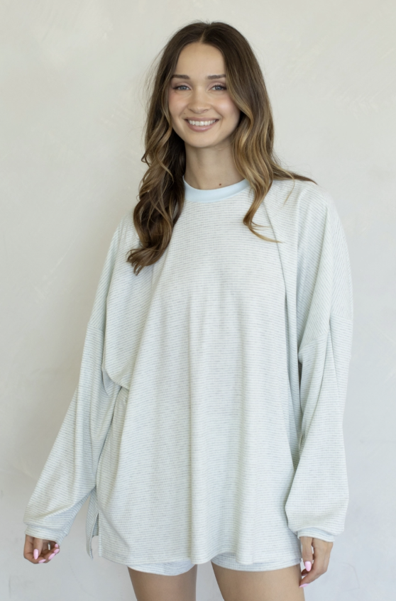 A woman wearing an oversized, light gray ribbed knit long-sleeve top with a crew neck, standing against a plain off-white wall.