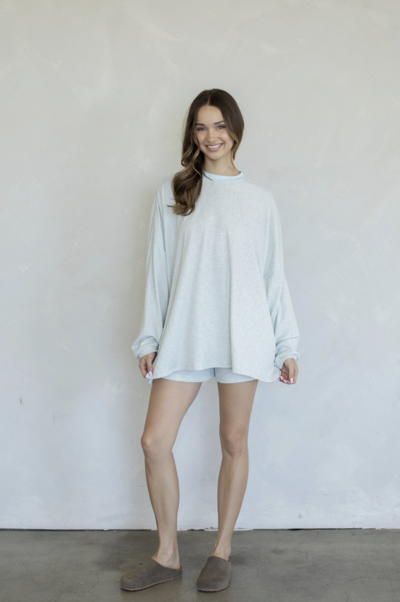 A woman modeling a light gray oversized long-sleeve top and matching shorts set, paired with taupe slip-on slippers, standing against a textured off-white wall.