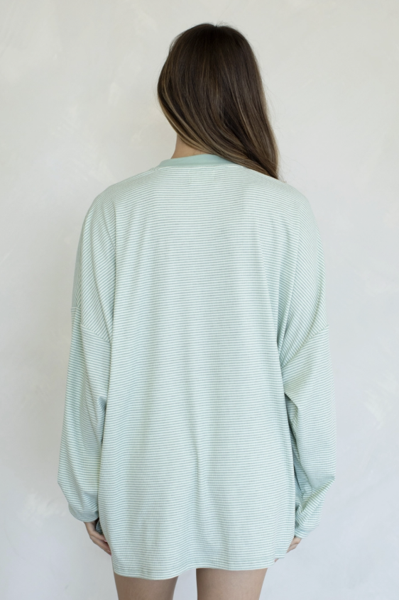 A woman viewed from behind wearing a pale mint green long-sleeve top with fine horizontal stripes, set against a plain off-white background.