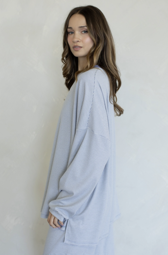 A woman with long brown hair is shown from the side/back wearing a loose, oversized long-sleeve top with fine horizontal blue and white stripes against a light textured wall.