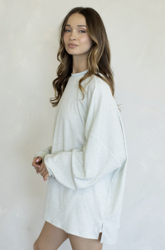 A woman wearing an oversized, long-sleeve ribbed knit sweater in a very light off-white or pale gray color, standing against a plain light background.