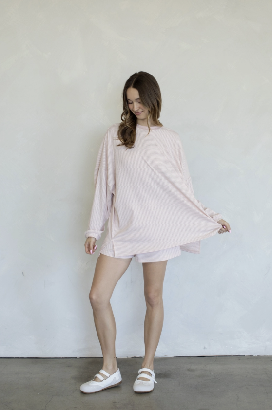 A woman modeling a light pink oversized long-sleeve tunic top and matching shorts set, paired with white flat shoes, standing against a textured off-white wall.