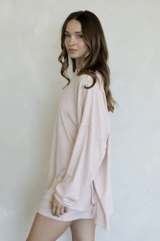 A woman wearing a light pink, oversized long-sleeve knit top with a subtle horizontal stripe texture, standing against a light gray textured wall.