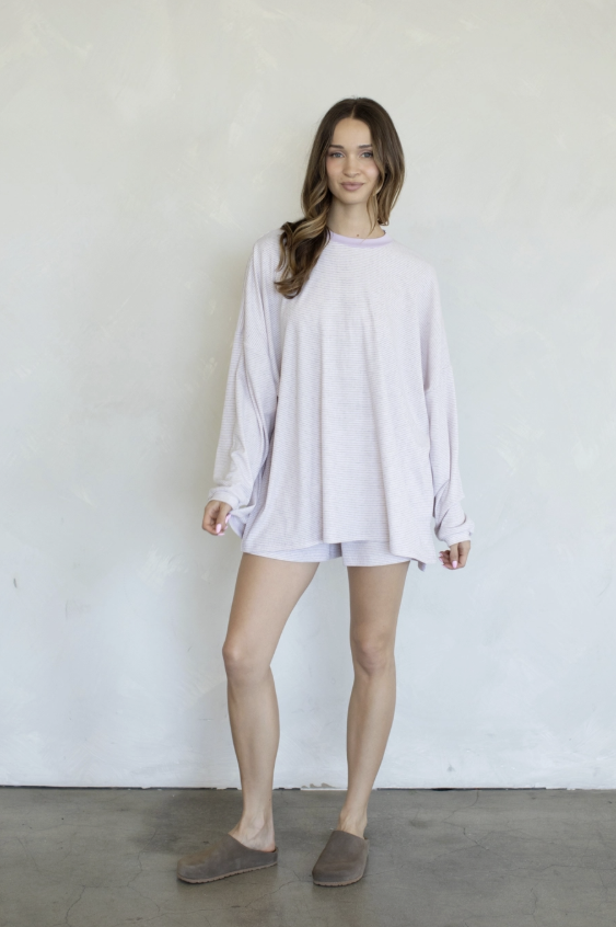 A woman modeling a light lavender oversized long-sleeve t-shirt dress with a crew neck, paired with brown slip-on slippers, standing against a textured off-white wall.