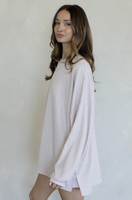 A woman wearing a loose, oversized long-sleeve top in a very pale pink or blush color, standing against a textured off-white wall.