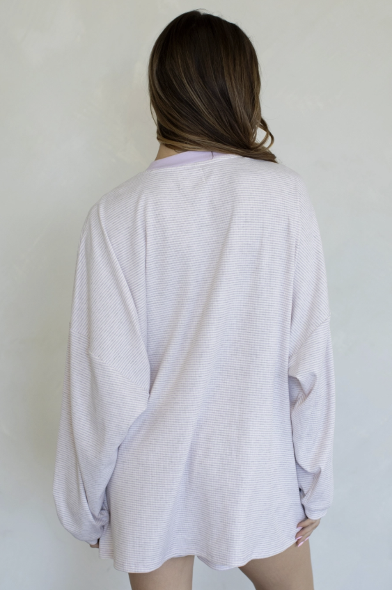 A woman viewed from behind wearing a light lavender long-sleeve knit top with a subtle horizontal ribbed texture, set against a plain off-white wall.