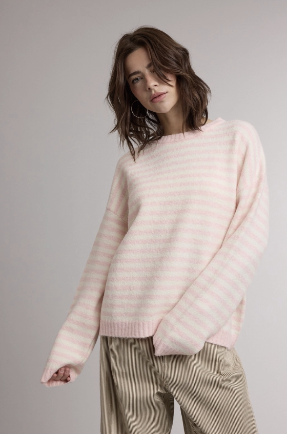 A woman wearing a light pink and white striped knit sweater with oversized sleeves, paired with beige corduroy trousers, photographed against a neutral gray studio background.