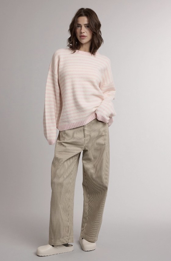 A woman modeling a light pink and white striped knit sweater paired with beige corduroy trousers and white slip-on shoes, photographed against a neutral light gray studio background.