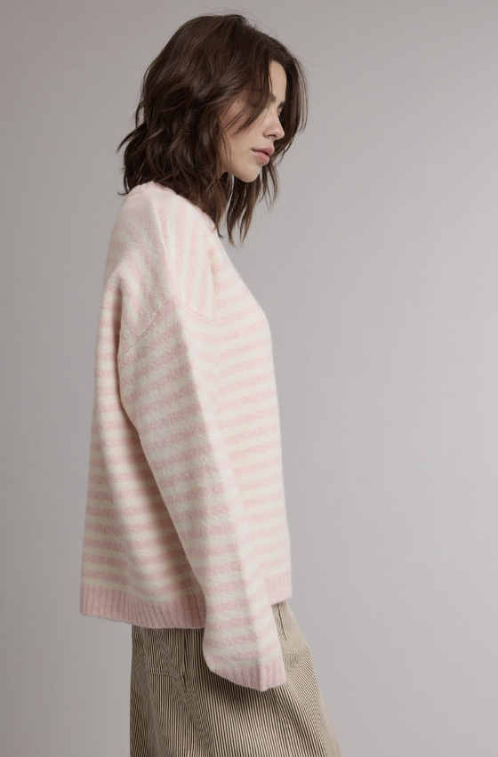 A woman shown in side profile wearing a soft knit sweater with horizontal light pink and off-white stripes, paired with beige corduroy trousers, against a plain light gray studio background.