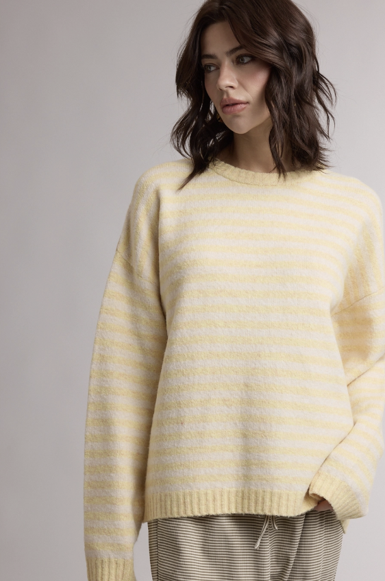 A woman wearing a cream and pale yellow striped knit sweater, paired with striped trousers, photographed against a neutral light gray studio background.
