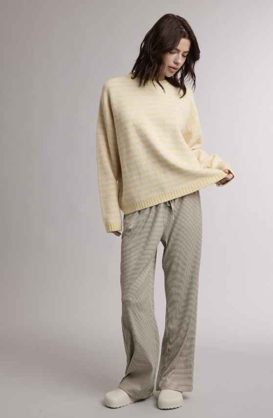 A woman modeling a cream-colored knit sweater and beige striped lounge pants against a neutral gray studio background.