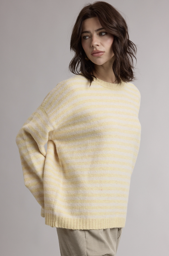 A woman wearing a cream and pale yellow striped knit sweater, shown from the waist up against a neutral gray studio background.