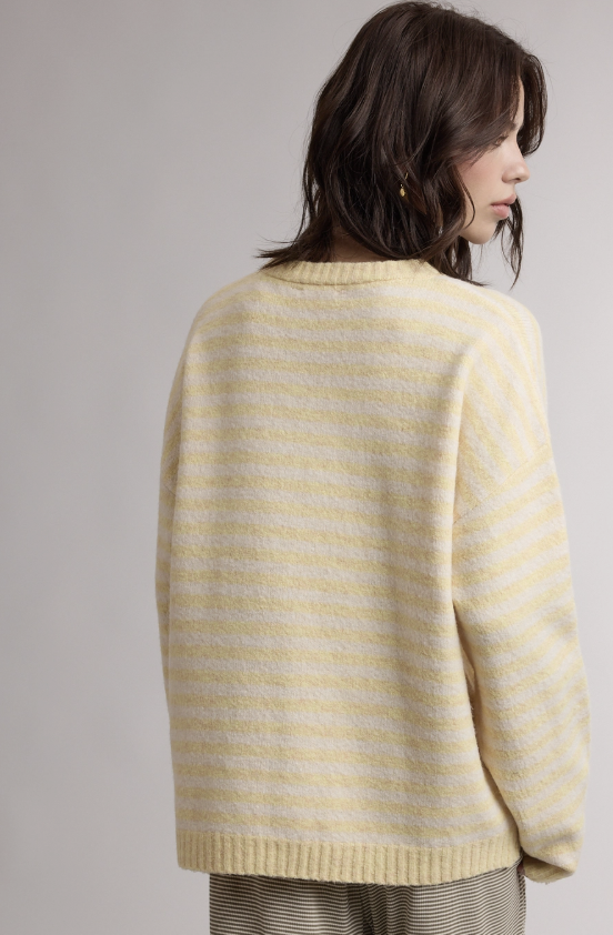 A woman shown from the back wearing a cream and pale yellow striped knit sweater, paired with striped trousers, against a neutral light gray background.