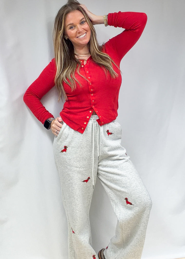 Woman wearing grey sweatpants with embroidered dogs in sweaters and santa hats with red top on plain background