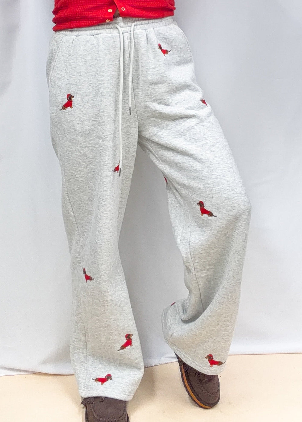 Woman wearing grey sweatpants with embroidered dogs in red sweaters and santa hats on plain background