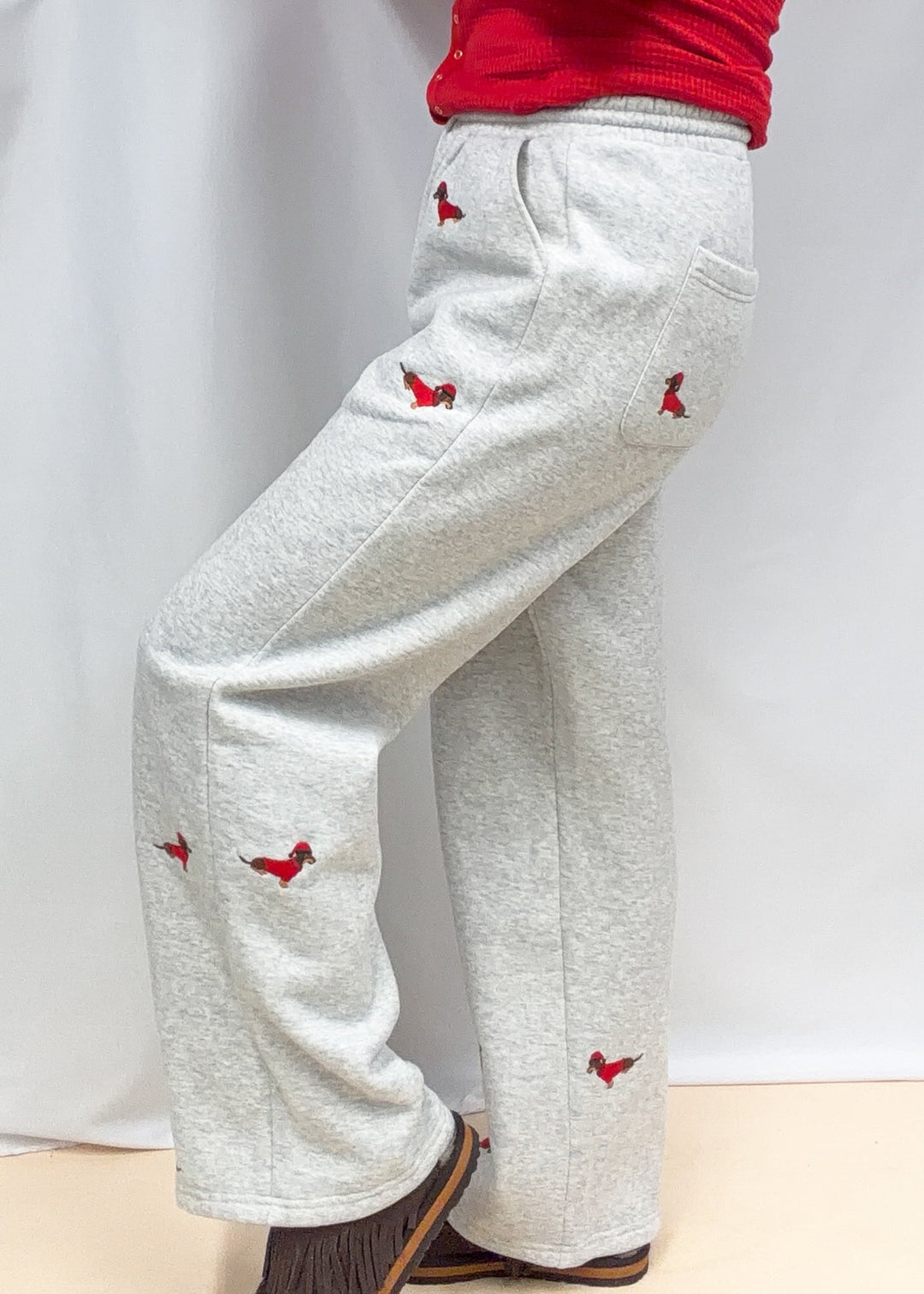 Woman shown from side view wearing grey sweatpants with embroidered dogs in red sweaters and santa hats on plain background
