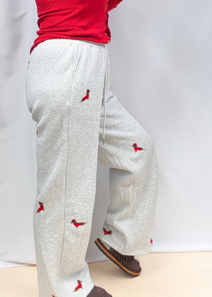 Woman wearing grey sweatpants with embroidered dogs in red sweater and santa hats on plain background