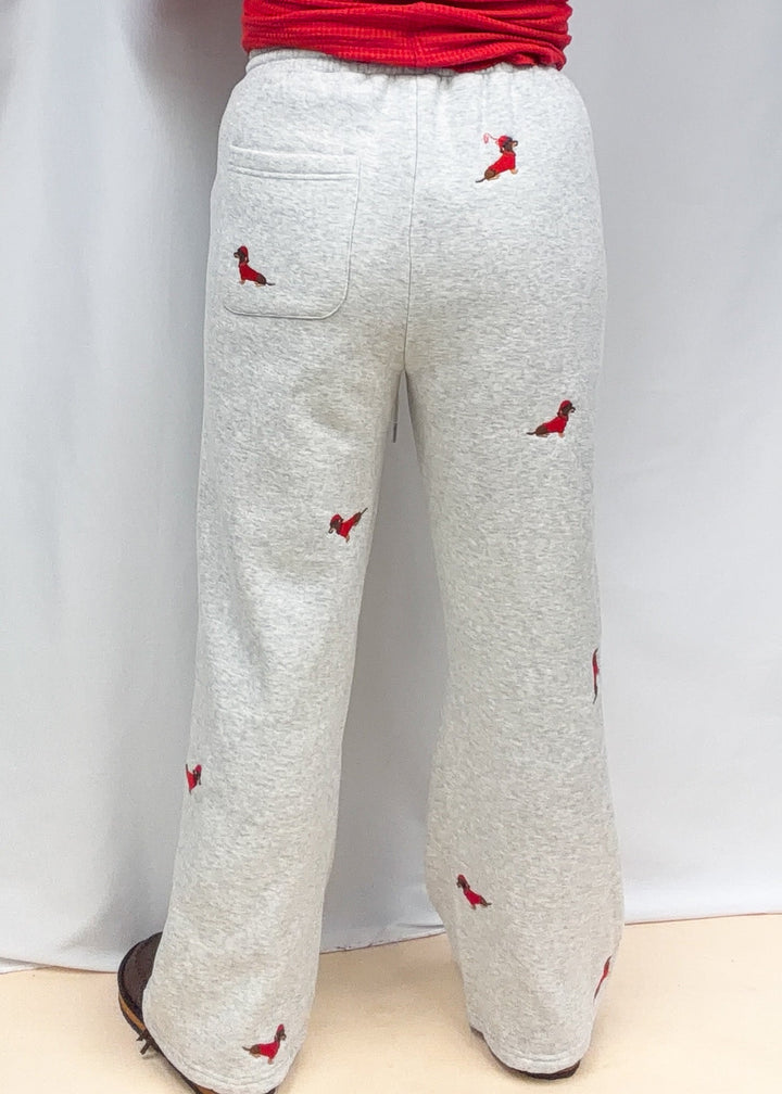 Woman shown from back view wearing grey sweatpants with embroidered dogs in red sweaters and santa hats on plain background