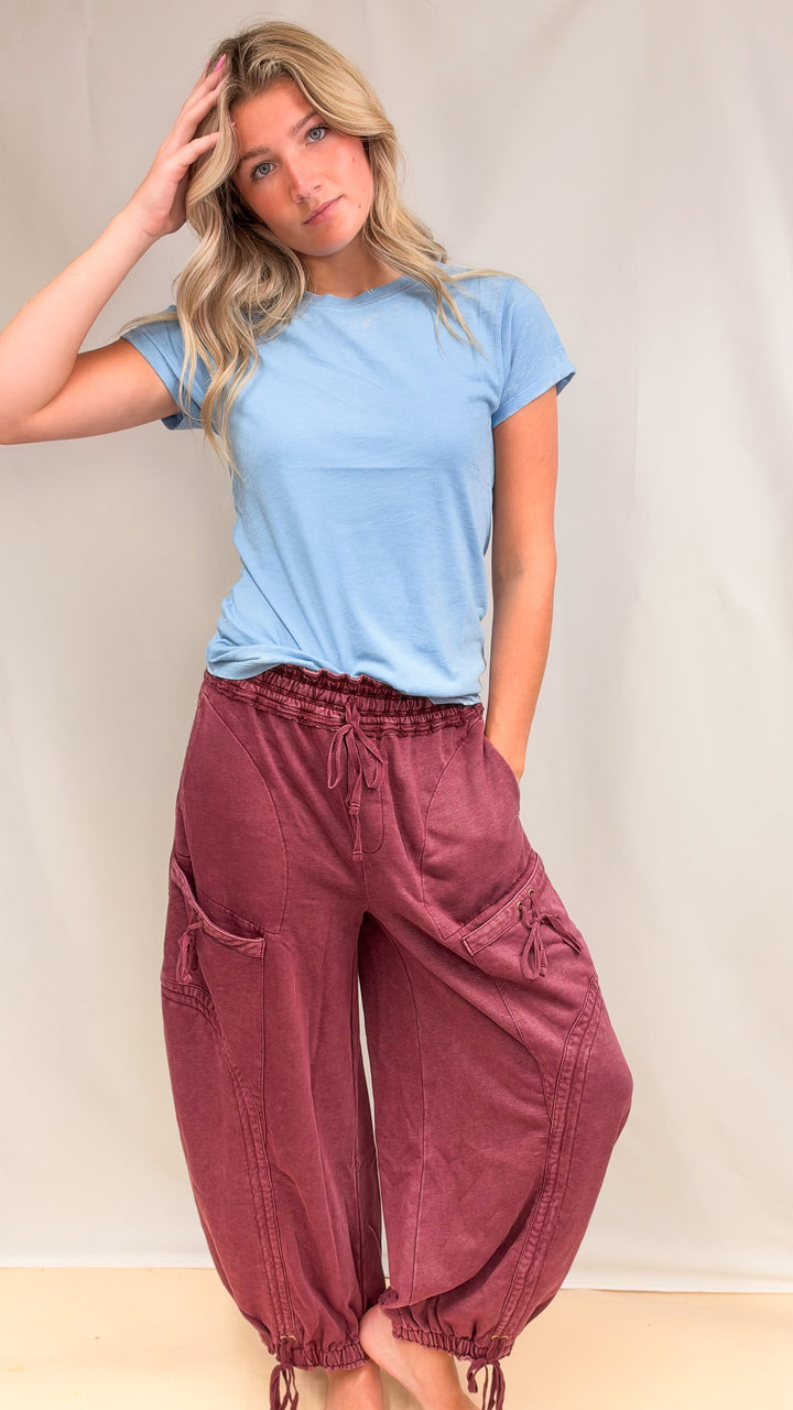 Woman wearing blue tee with maroon parachute pants with drawstring details on plain background