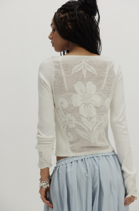 A woman shown from the back wearing a white long-sleeve crop top with a sheer floral lace back panel and a light blue skirt, against a plain light gray studio background.