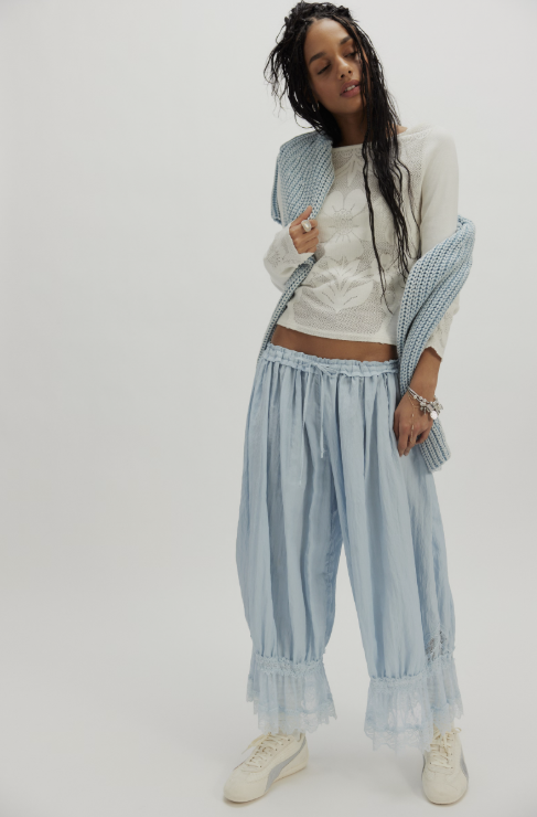 A woman modeling a light blue striped lounge set consisting of wide-leg pants with ruffled hems and a matching knit cardigan, styled with a white crop top and white sneakers against a plain light gray background.