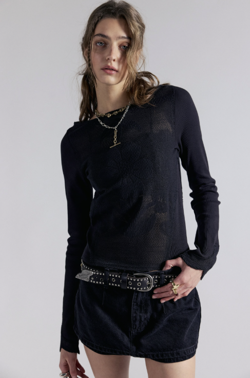 A model wearing a black long-sleeve knit top with a distressed texture, styled with dark denim shorts and a studded belt against a plain light gray background.