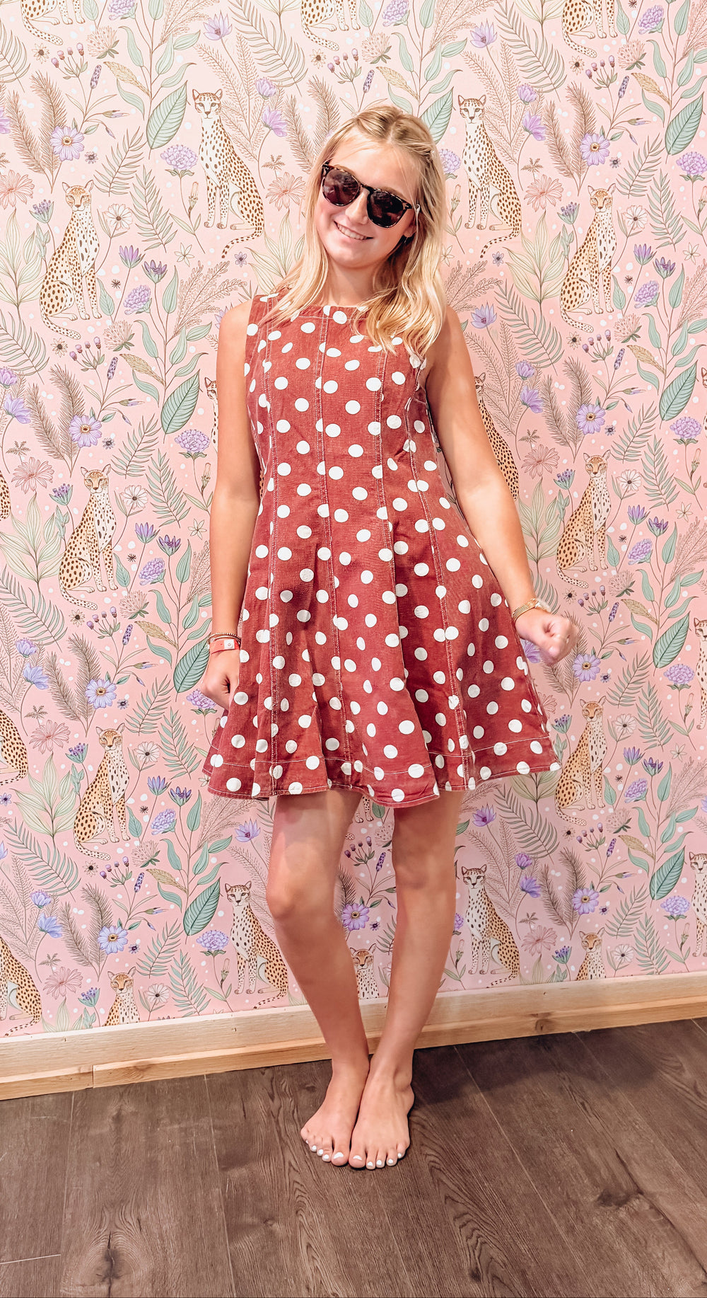 A young girl with blonde hair and sunglasses wearing a sleeveless rust-red dress with white polka dots, standing barefoot in front of a pink floral wallpaper background.