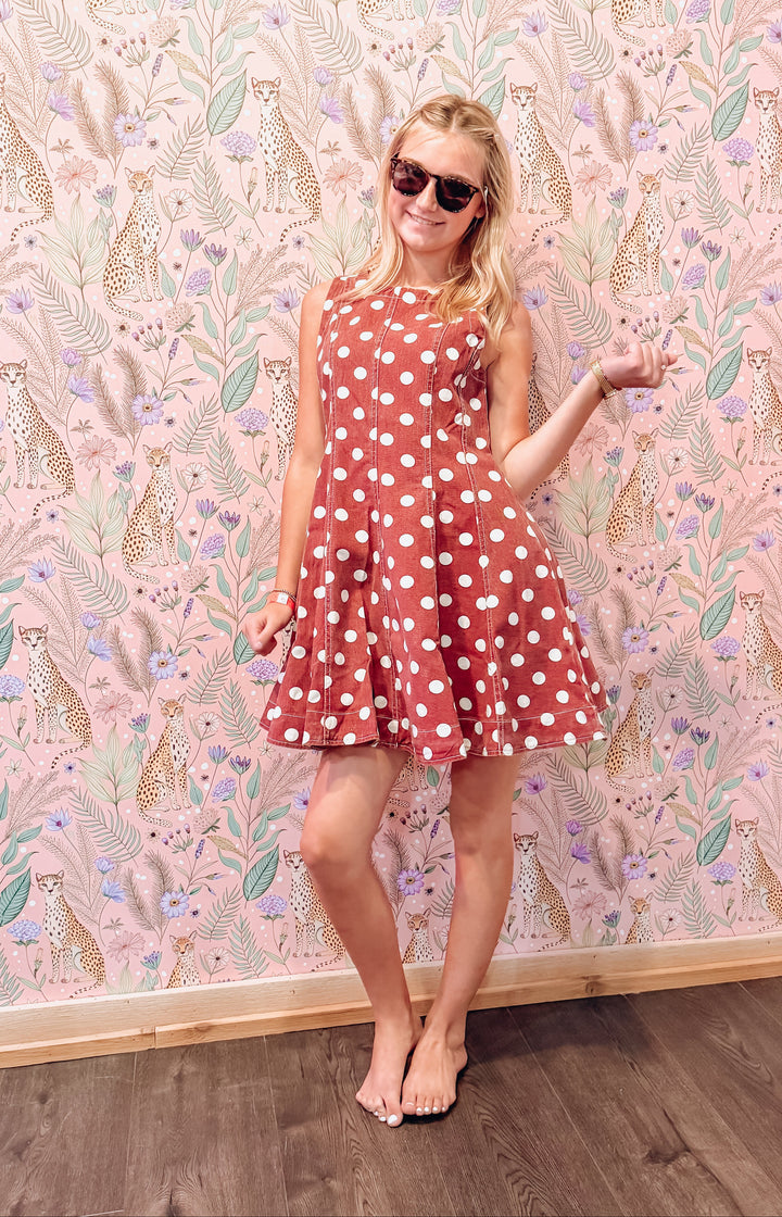 A woman wearing a sleeveless rust-red A-line dress with white polka dots and dark sunglasses, posing in front of a pink patterned wallpaper background.