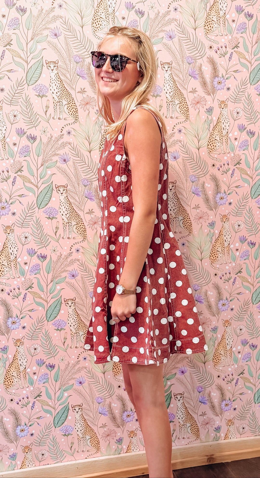A woman wearing a rust red sleeveless dress with white polka dots stands in front of a pink patterned wallpaper featuring leopards and foliage.