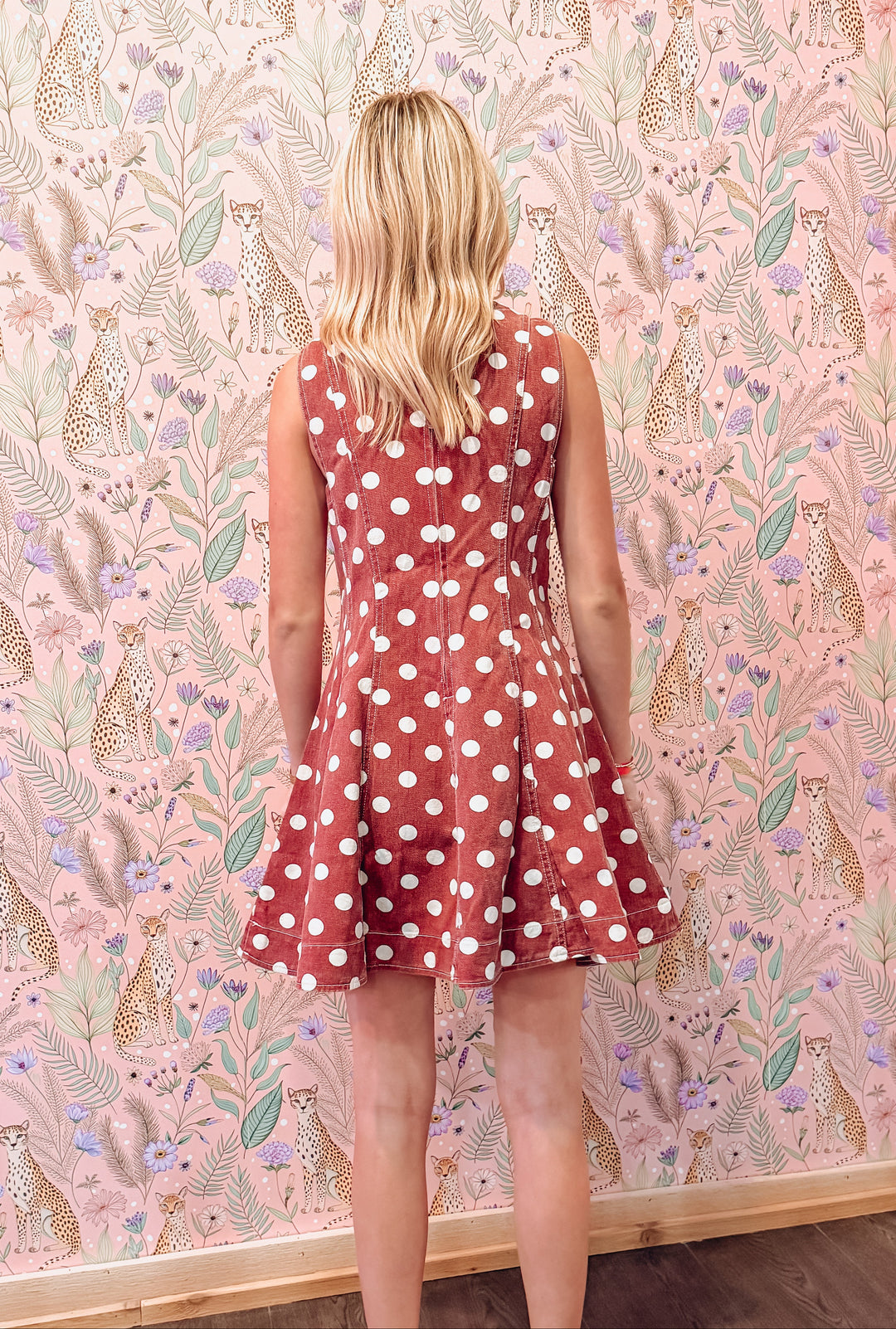 A woman with blonde hair is shown from behind wearing a sleeveless rust-red skater dress with white polka dots, standing in front of a pink patterned wallpaper featuring animals and leaves.