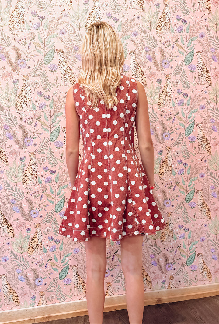 A woman with blonde hair is shown from behind wearing a sleeveless rust-red skater dress with white polka dots, standing in front of a pink patterned wallpaper featuring animals and leaves.