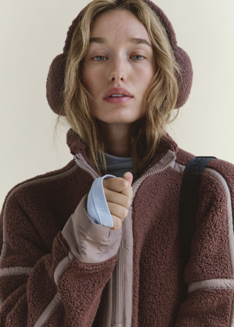 Woman wearing brown plush fleece ear warmers and brown fleece jacket on plain background