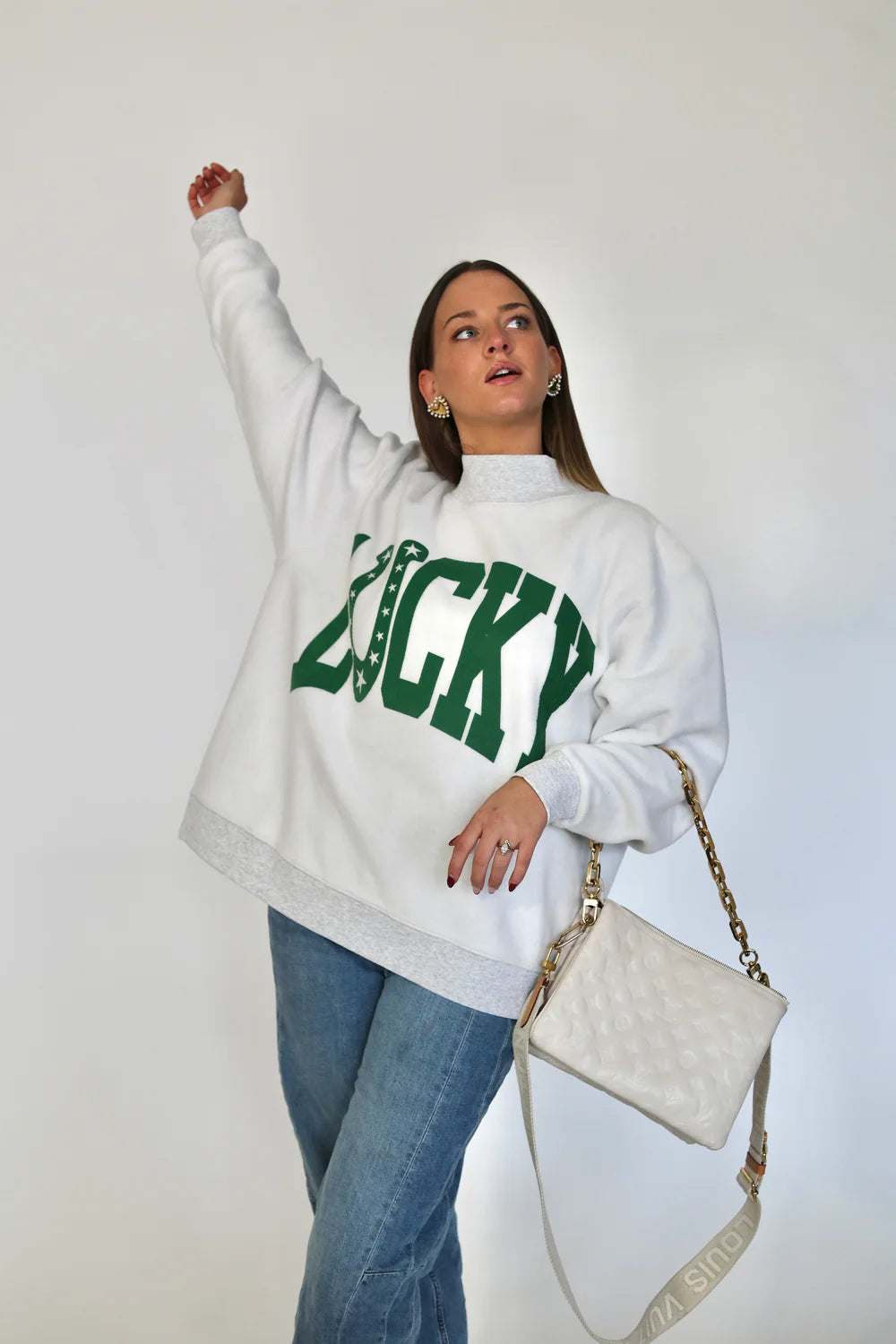 Woman wearing a white sweatshirt with green text, blue jeans, and holding a white handbag against a plain background.