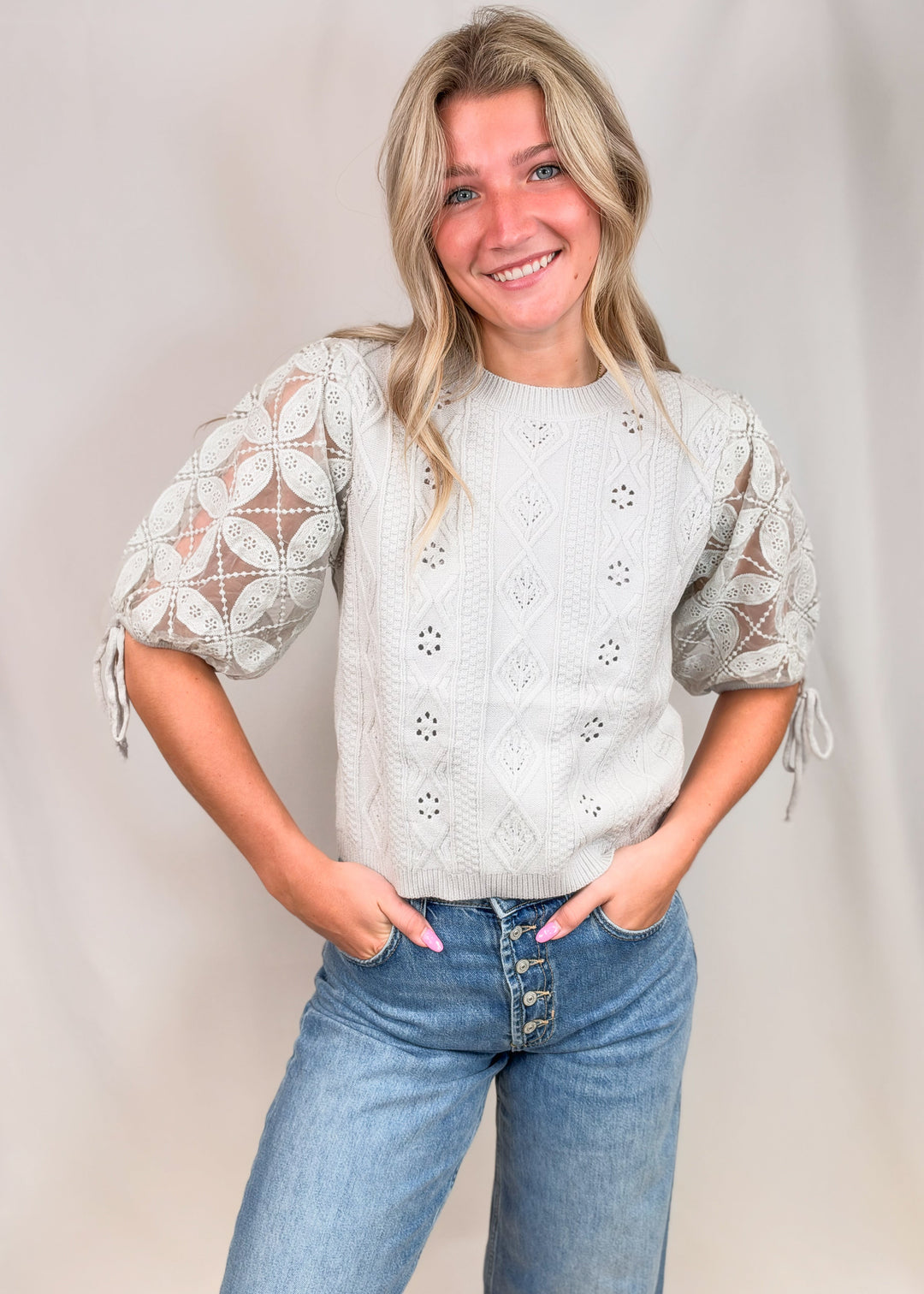 A smiling blonde woman wearing a light gray textured knit top with sheer lace puff sleeves and blue jeans, standing against a neutral light gray studio backdrop.