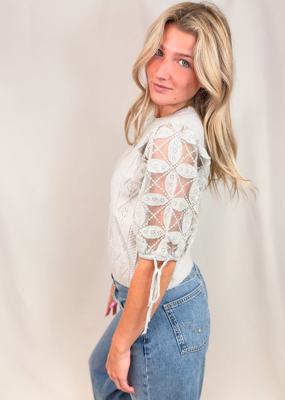 A blonde woman wearing a white crochet lace top with sheer sleeves and blue jeans, posing against a light beige studio backdrop.