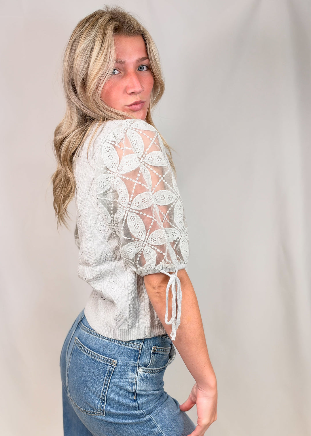 A woman wearing a white crochet lace top with tie details on the sleeves and blue jeans, posing against a light beige studio backdrop.