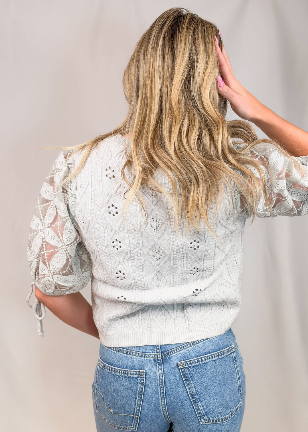 A woman shown from the back wearing a light gray eyelet lace knit sweater with sheer puff sleeves and blue jeans, standing against a neutral light gray studio backdrop.