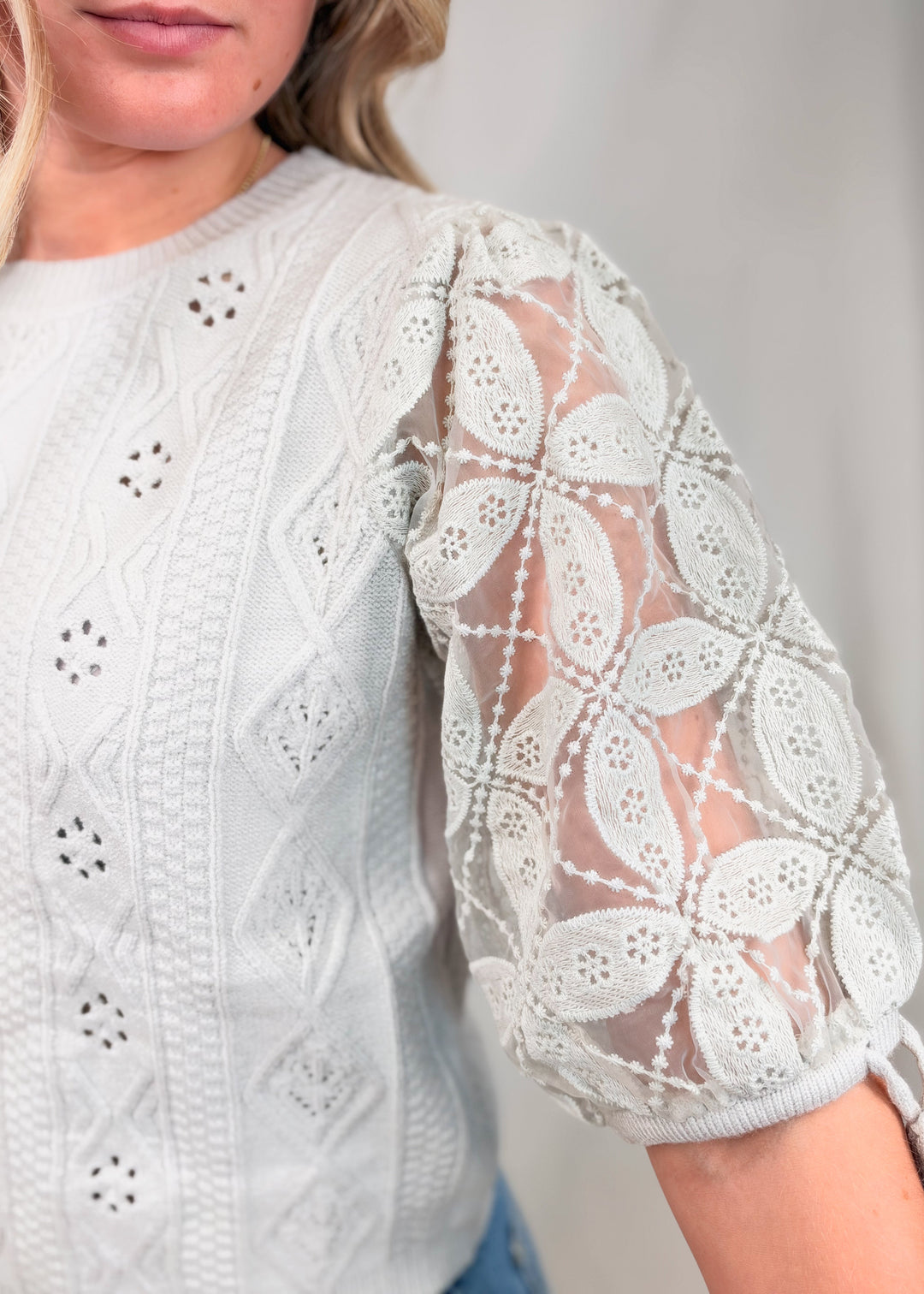 Close-up of a woman wearing a white eyelet knit top with sheer floral lace puff sleeves and side tie details.