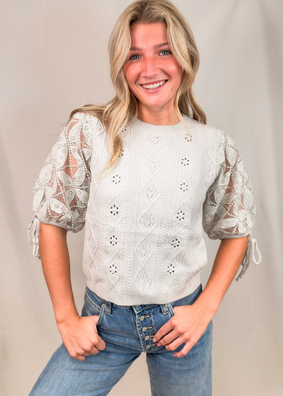 A smiling blonde woman wearing a cream-colored eyelet knit sweater with sheer lace puff sleeves and tie details, paired with blue jeans, standing against a neutral beige studio backdrop.
