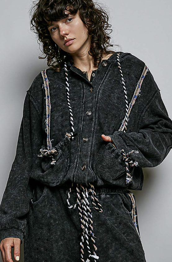 A model wearing a dark washed black corduroy button-up shirt jacket with decorative braided rope details down the front and on the sleeves, paired with a matching dark corduroy skirt against a plain gray studio background.