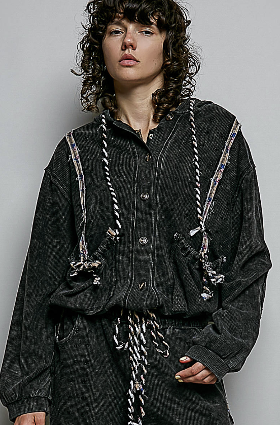 A model with curly dark hair wearing a dark washed black hooded button-up shirt with decorative braided rope details and a matching dark textured bottom, photographed against a plain light gray studio background.