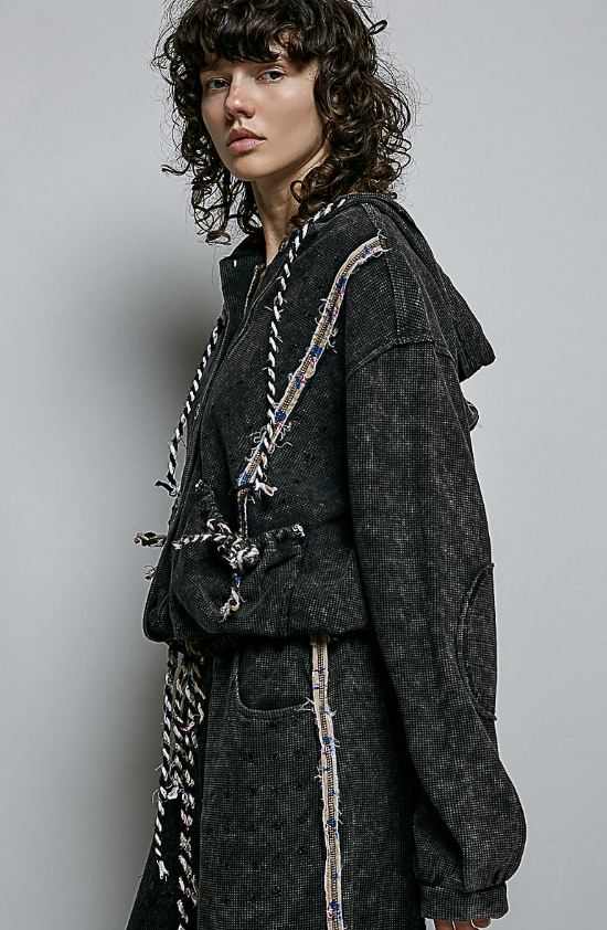 A model wearing a dark gray acid-washed hooded jacket with unique braided rope details and a matching dark skirt, photographed against a neutral gray background.