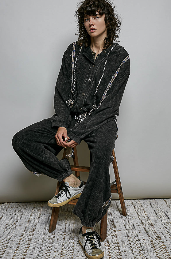 A model with curly dark hair sits on a wooden stool wearing a matching black washed corduroy shirt and wide-leg pants set, styled with distressed white and black sneakers against a neutral gray background.