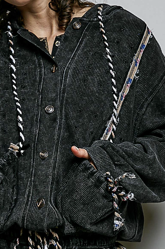 Close-up of a person wearing a dark charcoal tweed button-front jacket with braided trim details and a patterned strap across the chest.