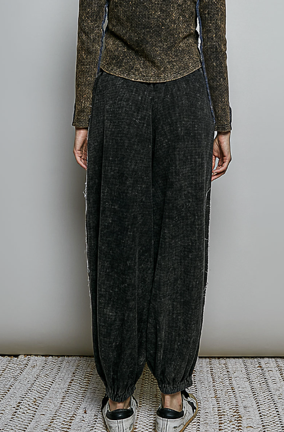 A person shown from the chest down wearing dark charcoal gray textured wide-leg pants with side slits, a brown textured long-sleeve top, and white sneakers, standing against a plain light gray wall.