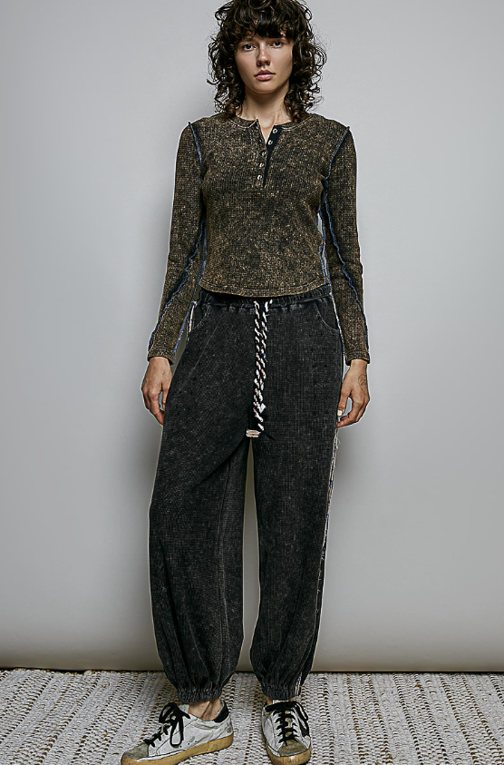 A model stands against a light gray wall wearing a brown and black textured long-sleeve henley top with dark charcoal jogger pants featuring a white braided drawstring, and white sneakers with gum soles.