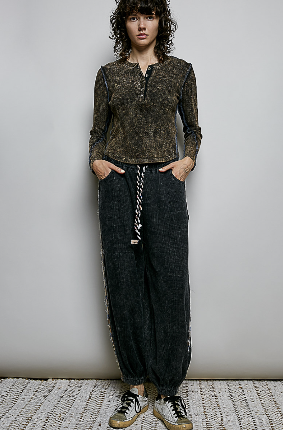 A model stands against a plain light gray wall wearing a dark brown marled long-sleeve henley top, dark charcoal gray textured jogger pants with a black and white braided drawstring, and distressed sneakers.